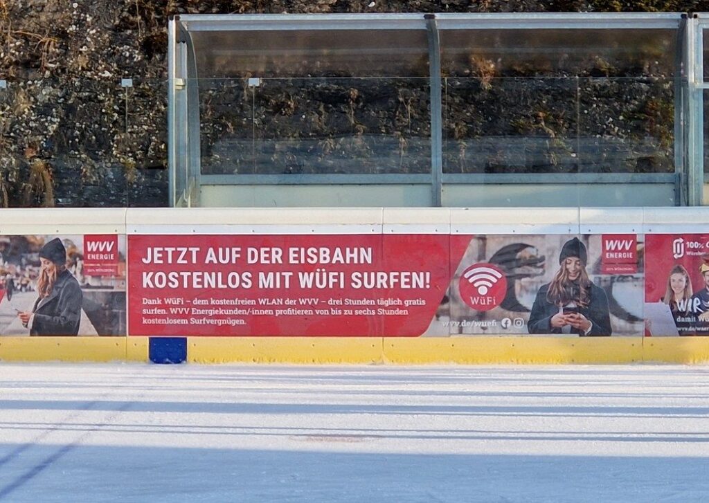 Bande der Eisbahn Würzburg mit einem Hinweis auf das kostenlose WLAN WüFi der WVV, davor die Eisfläche