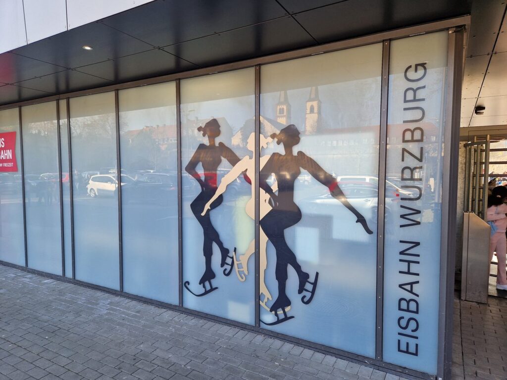 Glasfassade am Eingang der Eisbahn Würzburg mit Silhouetten von drei Eiskunstläuferinnen und dem Schriftzug Eisbahn Würzburg