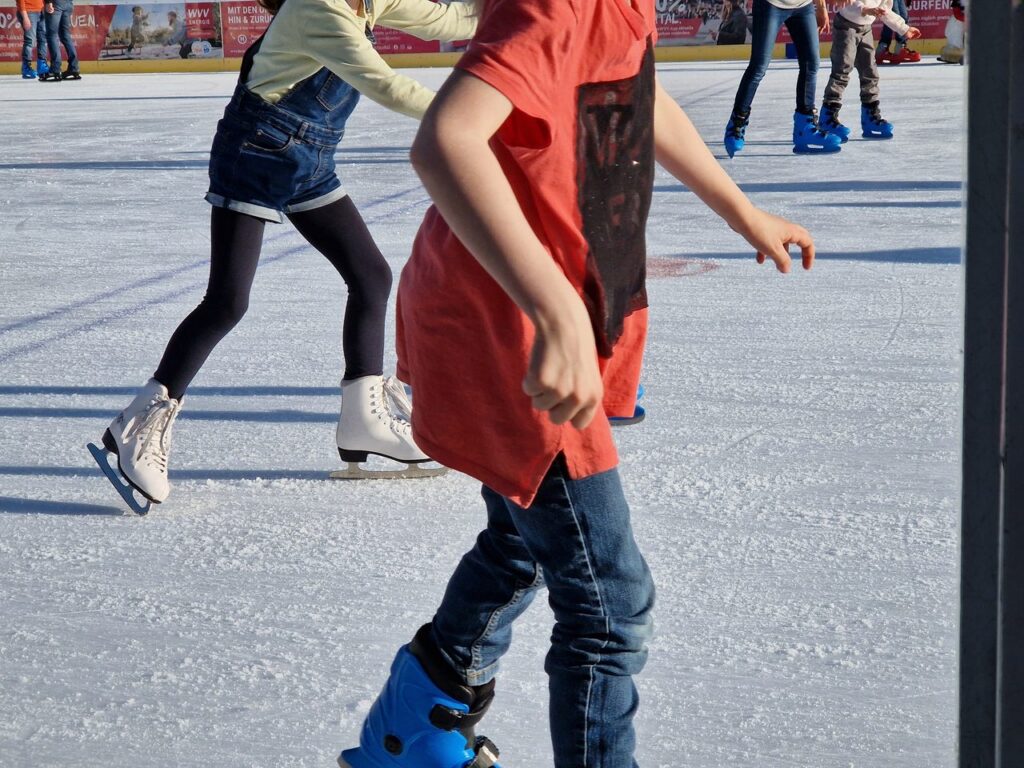 Kinder laufen auf der Eisfläche der Eisbahn Würzburg Schlittschuh, im Vordergrund Leihschlittschuhe in Blau, links ein weißer Eiskunstlaufschuh