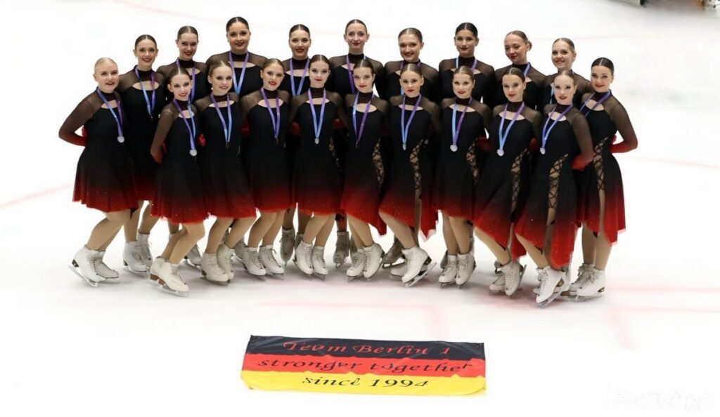 Gruppenfoto von Team Berlin 1 in schwarz-roten Kleidern mit Medaillen auf dem Eis, davor ein Banner mit der Aufschrift Team Berlin 1, stronger together, since 1994