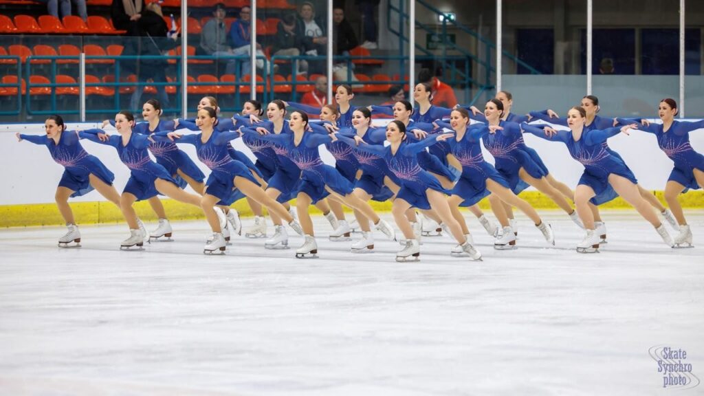 Synchroneiskunstlauf-Team der United Angels Stuttgart in blauen Kleidern in einer Linienformation mit ausgestreckten Armen auf dem Eis